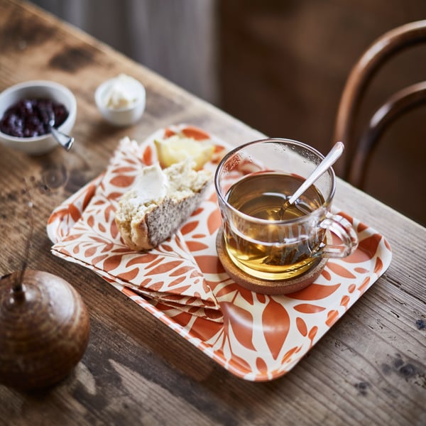 Holztisch mit HÖSTAGILLE Tablett im orange-weißen Blättermuster, darauf ein Glas Tee mit Löffel, Brot mit Käse, eine kleine Schale Oliven und ein Holzbehälter daneben.