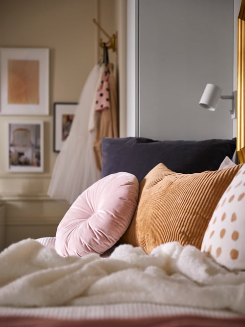 A round light pink KRANSBORRE cushion and various other cushions on a daybed with a white throw, beside a white wardrobe.