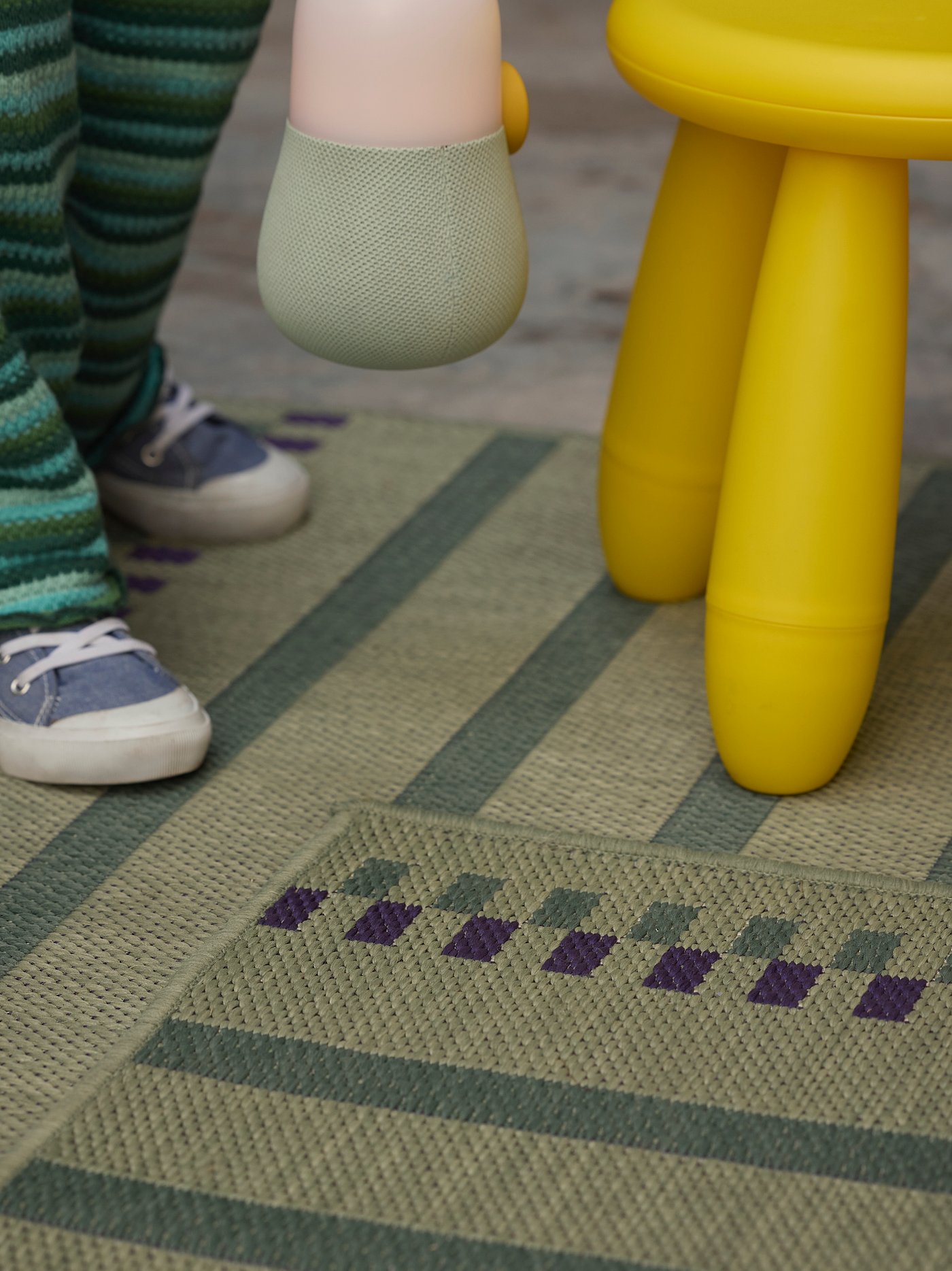 Primer plano de una alfombra plana KORSNING a rayas verdes y moradas, dos patas visibles de un taburete amarillo y los pies de una persona.