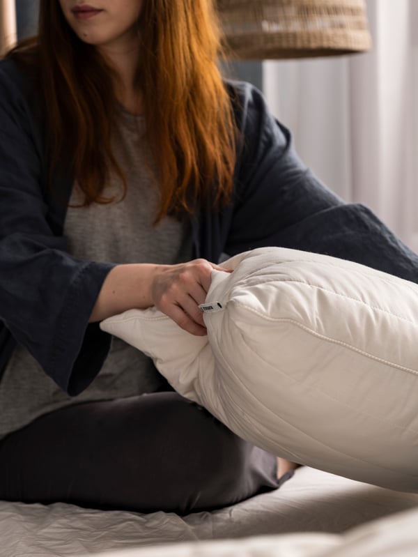 A person in a dark dressing gown is sitting on a bed and lifting a RUMSMALVA ergonomic pillow for side or back sleepers.