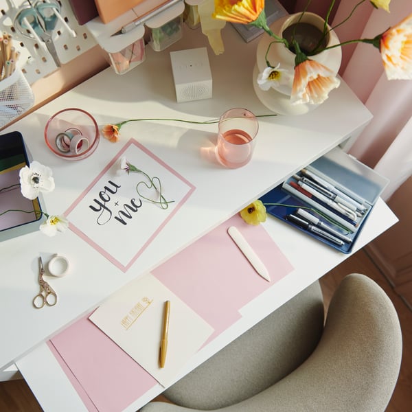 A dark beige KRYLBO swivel chair stands at a white LÄNGDHOPP pull-out desk with many items on top of it.
