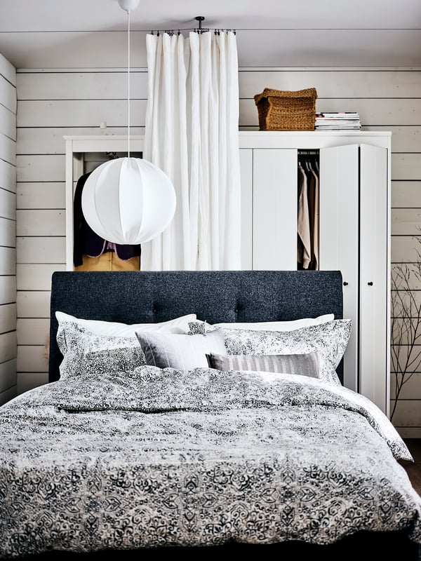 A dark grey IDANÄS upholstered bed covered with ÄNGSKLOCKA bed linen in front of a white curtain and two wardrobes.