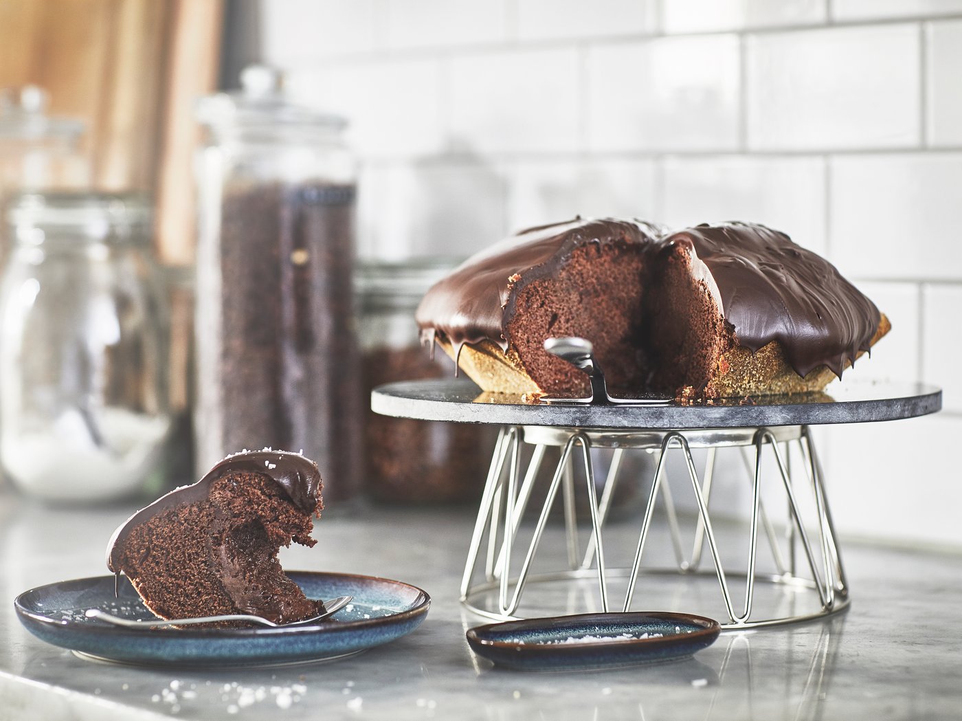 En chokoladekage placeret oven på et BAKGLAD kagefad. Et stykke kage ligger på en tallerken ved siden af fadet.