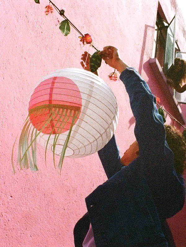 Una persona decora un espacio exterior con una lámpara colgante ÖMSESIDIG redonda y una guirnalda artificial con flores y hojas.