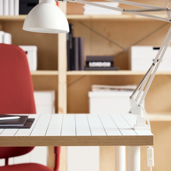 A close-up of a white/anthracite LAGKAPTEN tabletop with a TERTIAL work lamp attached and a red office chair in the back.