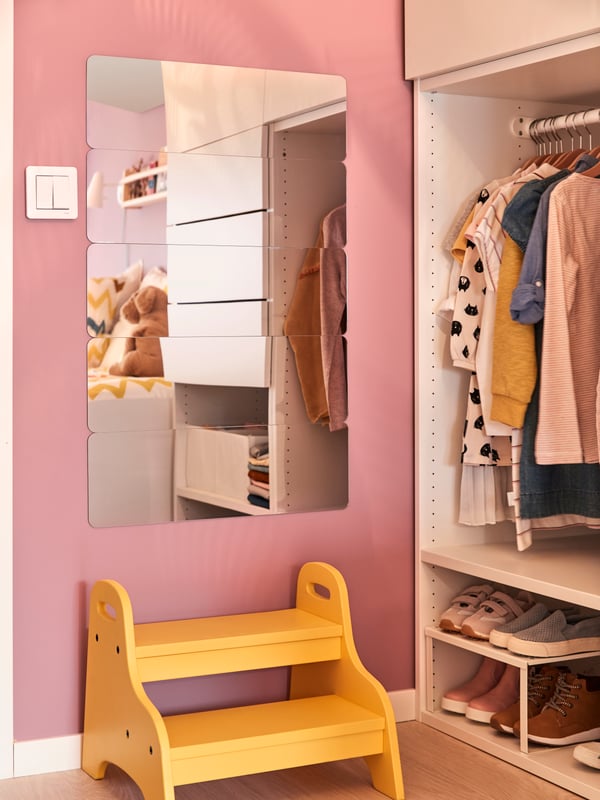 Un piso escalonado TROGEN amarillo situado frente a un espejo GOSIG GOLDEN en una habitación infantil con muros rosados y piso de madera. A un costado se ve un clóset PLATSA con ropa infantil colgada y zapatos ordenados con organizadores en la parte inferior. 