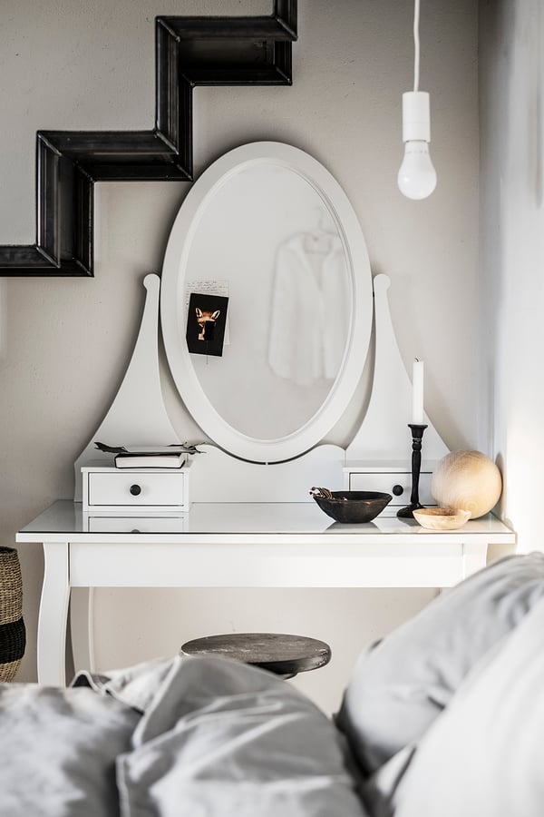 Coiffeuse HEMNES avec un plateau en verre et deux petits tiroirs et un miroir sur lequel se trouve un petit autocollant noir. Un petit tabouret en dessous de la coiffeuse avec un lit derrière et une lampe suspendu 