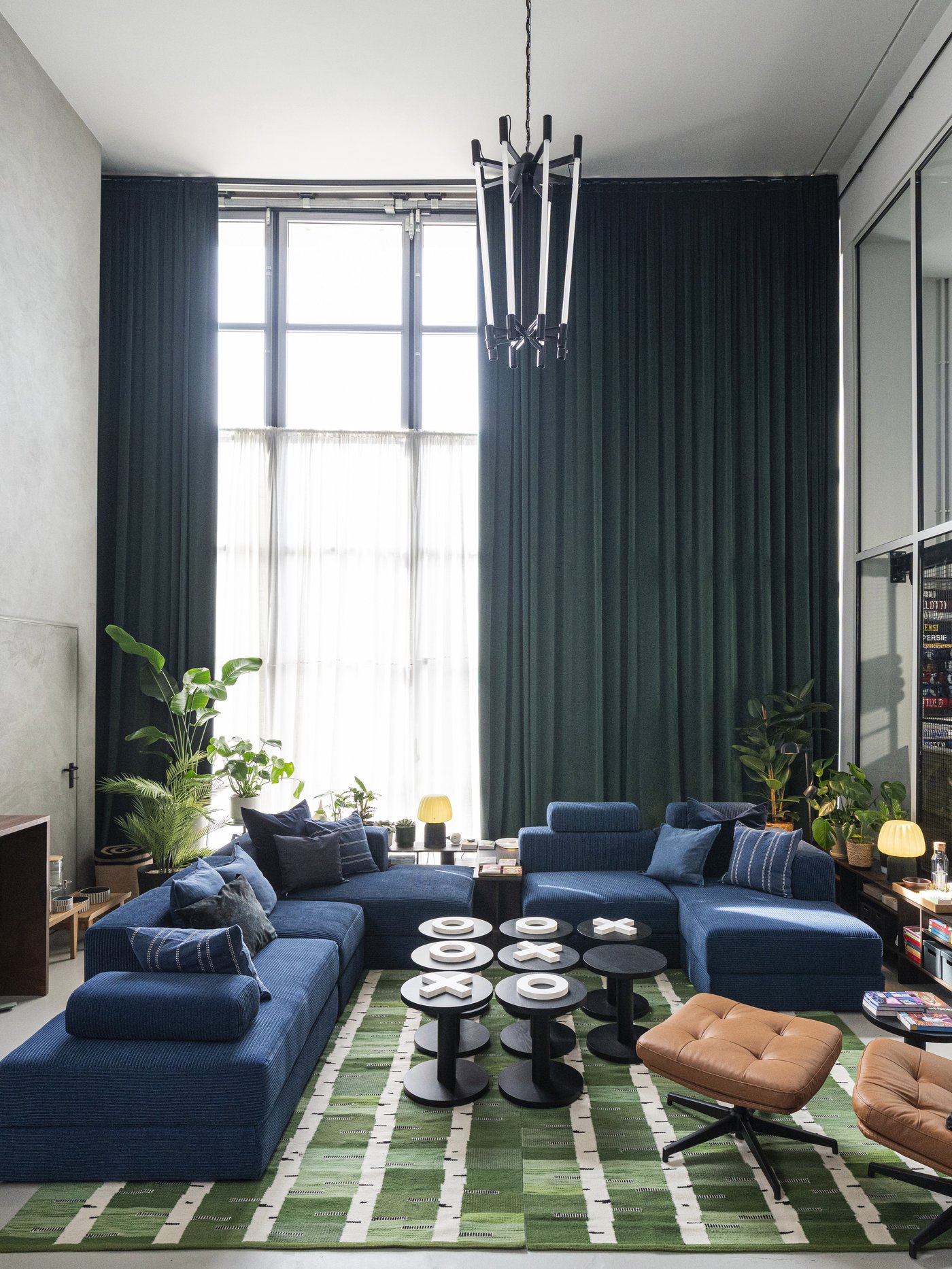 Ampio studio luminoso con grandi finestre, tende verde scuro e un divano modulare in velluto a coste blu JÄTTEBO. Al centro tavolini che formano un grande tris, sulla destra una poltrona con poggiapiedi color cuoio HAVBERG.