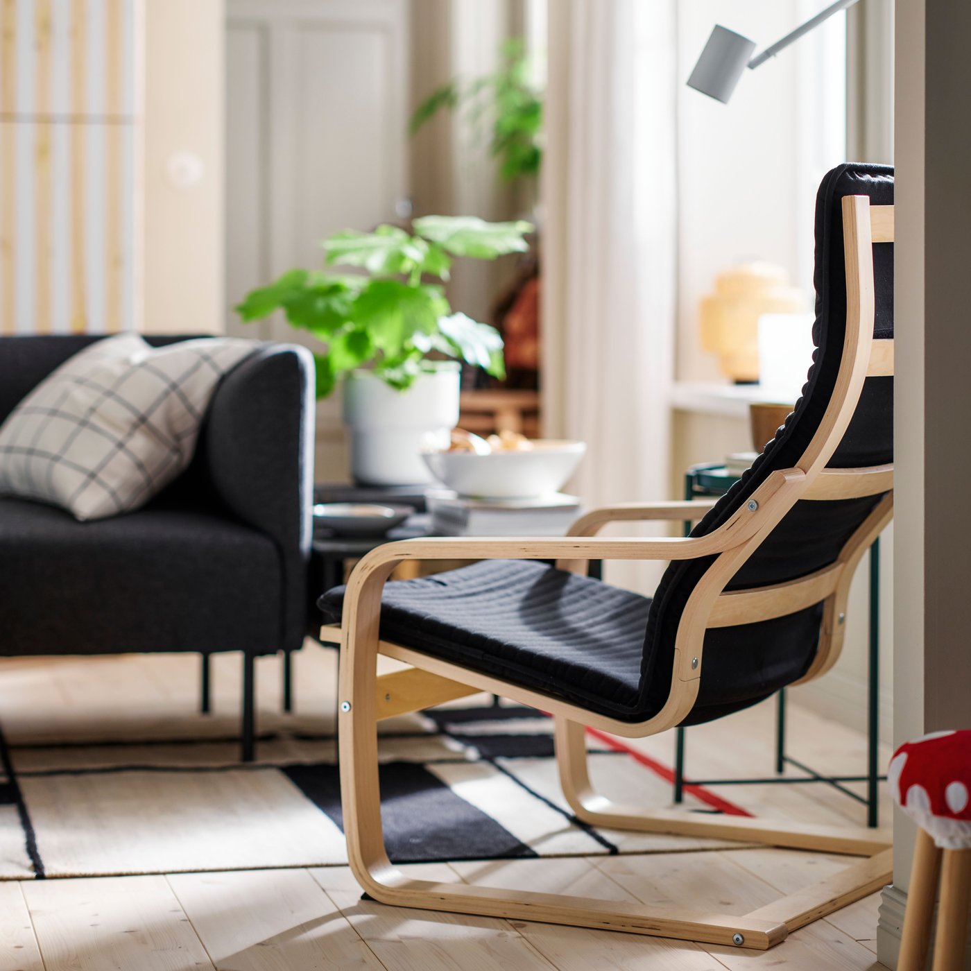 A bright living room with leafy plants and a black POÄNG armchair made with birch veneer, placed opposite a LILLEHEM sofa.