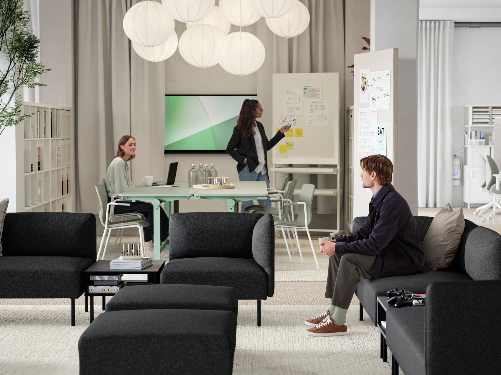 A young man sitting in a lounge area of several dark grey LILLEHEM sofa modules, TUNSTA tables and a meeting area behind it.