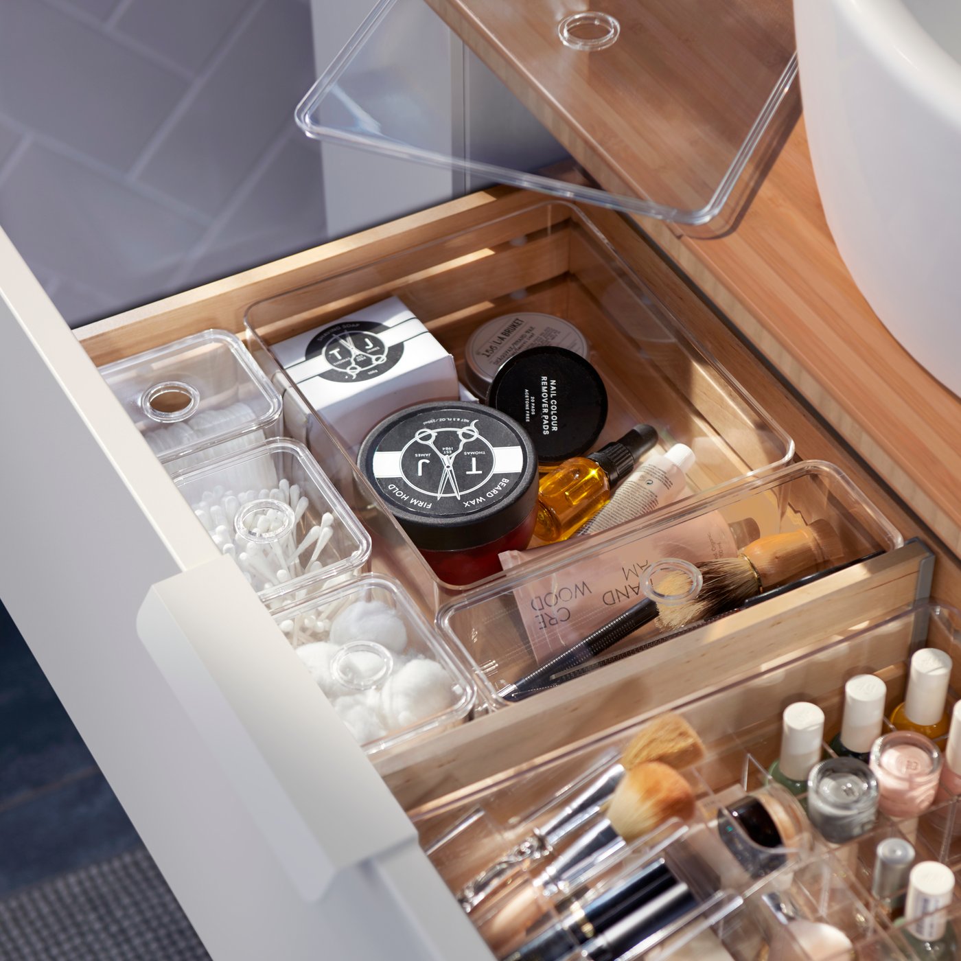 Eine Kombination aus mehreren GODMORGON Kästen mit Deckeln, in denen sich in einer holzfarbenen Schublade Badezimmerutensilien befinden.