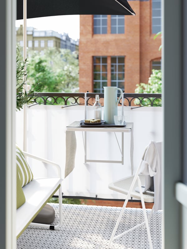 A small white balcony table hung on the balcony rail, with a pitcher, a plate of food and drinks on it.