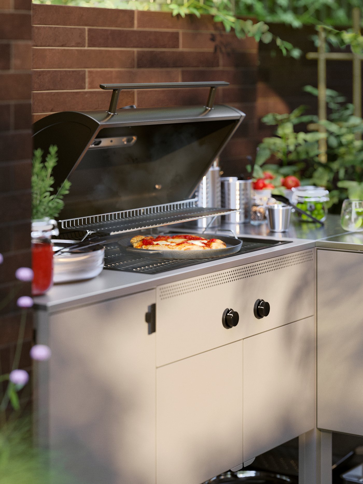 Un patio iluminado por la luz del atardecer y una pizza horneándose en una cocina exterior BÅTSKÄR con barbacoa de gas junto a hierbas aromáticas y tomates.