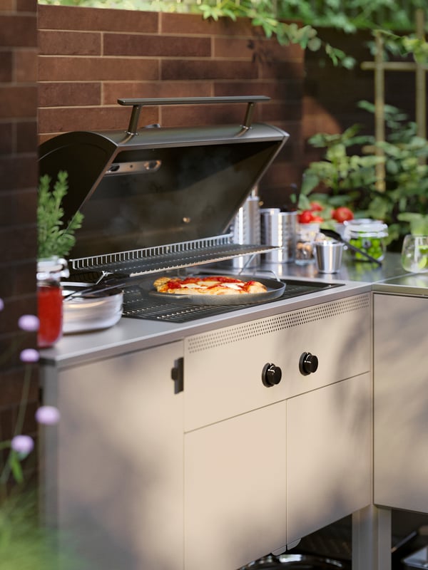 O terasă în lumina serii și o pizza care se coace într-o bucătărie de exterior BÅTSKÄR cu grătar pe gaz, lângă ierburi aromatice și roșii.