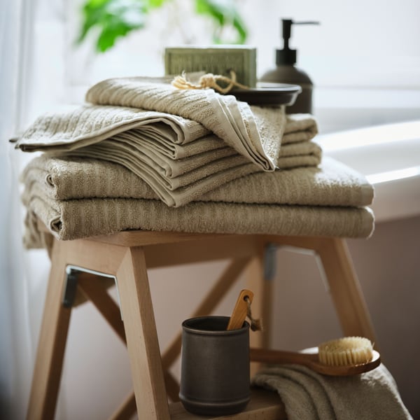 A pile of VÅGSJÖN cotton towels in beige colour on a wooden stool in front of a window. Soap and a body brush next to it.