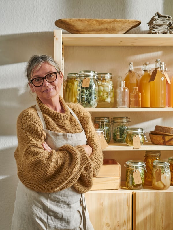 IKEA IVAR pine shelving unit with cabinet full of KORKEN jars and a KNAGGLIG box to store food. A lady wearing a working apron stand in front of the IVAR unit. 
