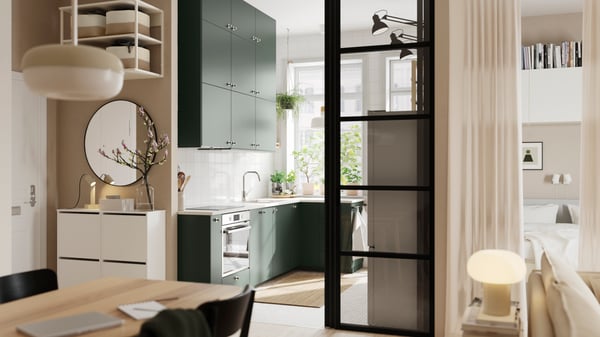 An open kitchen with deep green HAVSTORP kitchen fronts and wall cabinets up to the ceiling, next to a bright living room.