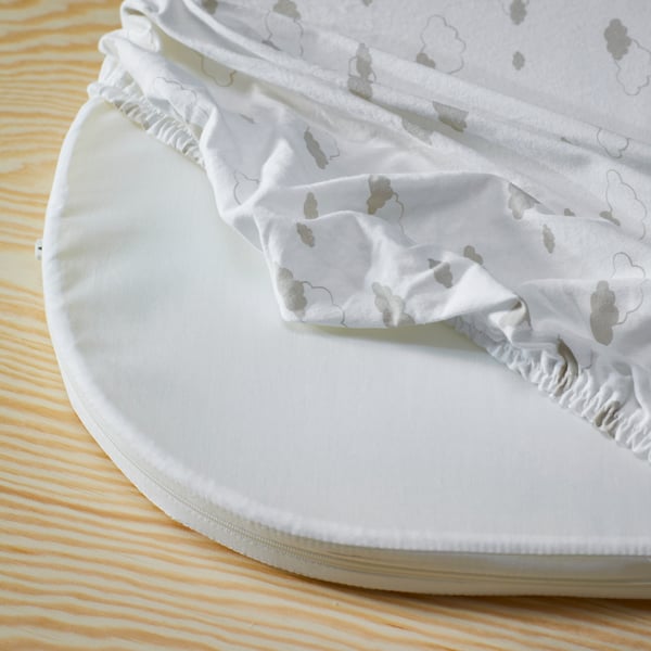 An ÄLSKVÄRD mattress for bassinet is being fitted with an ÄLSKVÄRD fitted sheet with clouds printed on it.