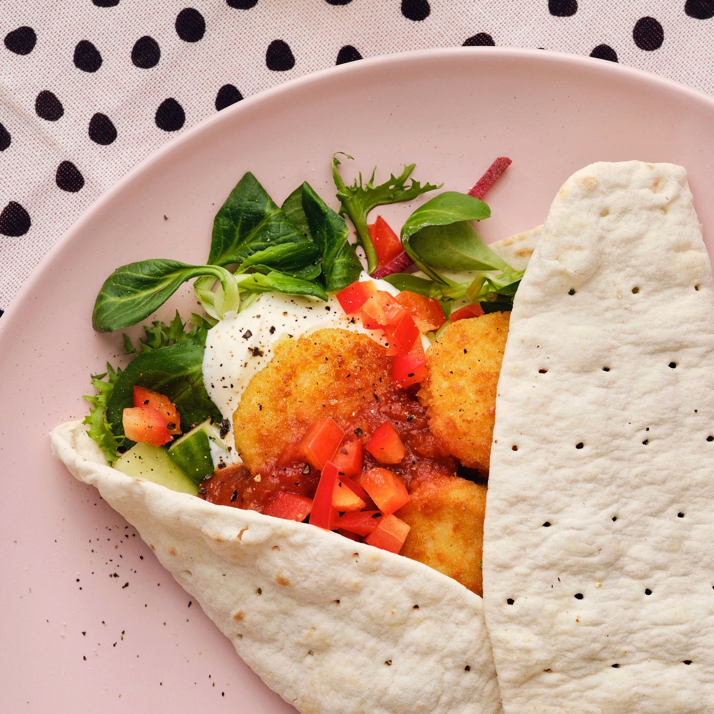 A pink plate with a wrap filled with lettuce, tomatoes, and SLAGVERK breaded wheat-based pieces.