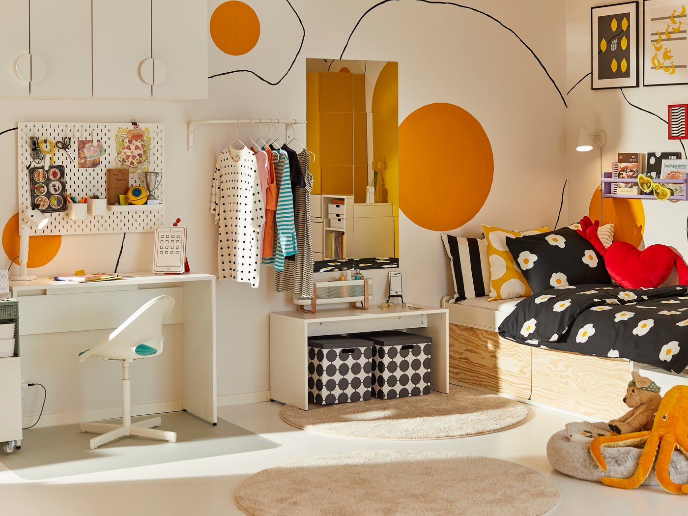 Светлая, современная спальня с белым письменным столом, яркой вешалкой для одежды и забавными оранжевыми кругами на стене. На кровати черно-белое одеяло с ромашками и подушки в виде красных сердечек. Корзины для хранения и круглый коврик дополняют уютное пространство.
