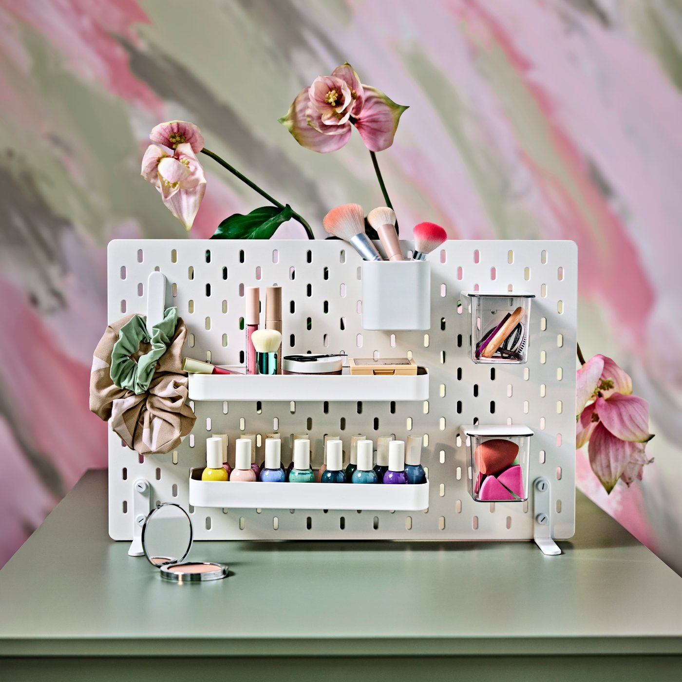 A white, freestanding SKÅDIS displaying nail polish and makeup, on a green table with pink flowers in the background.