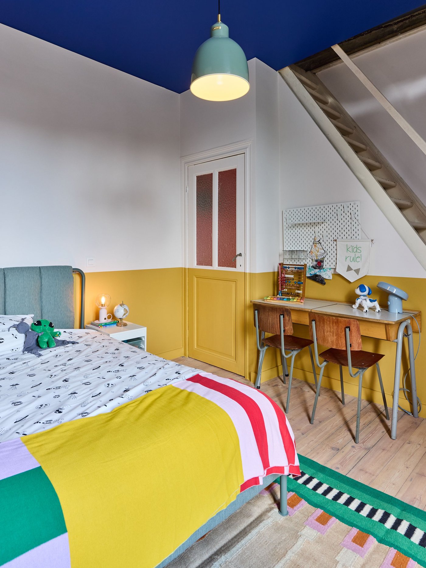 Slaapkamer met geel‑witte muur, bed met kleurrijke sprei, bureau met stoelen en trap naar boven, houten vloer en speelse, moderne inrichting.