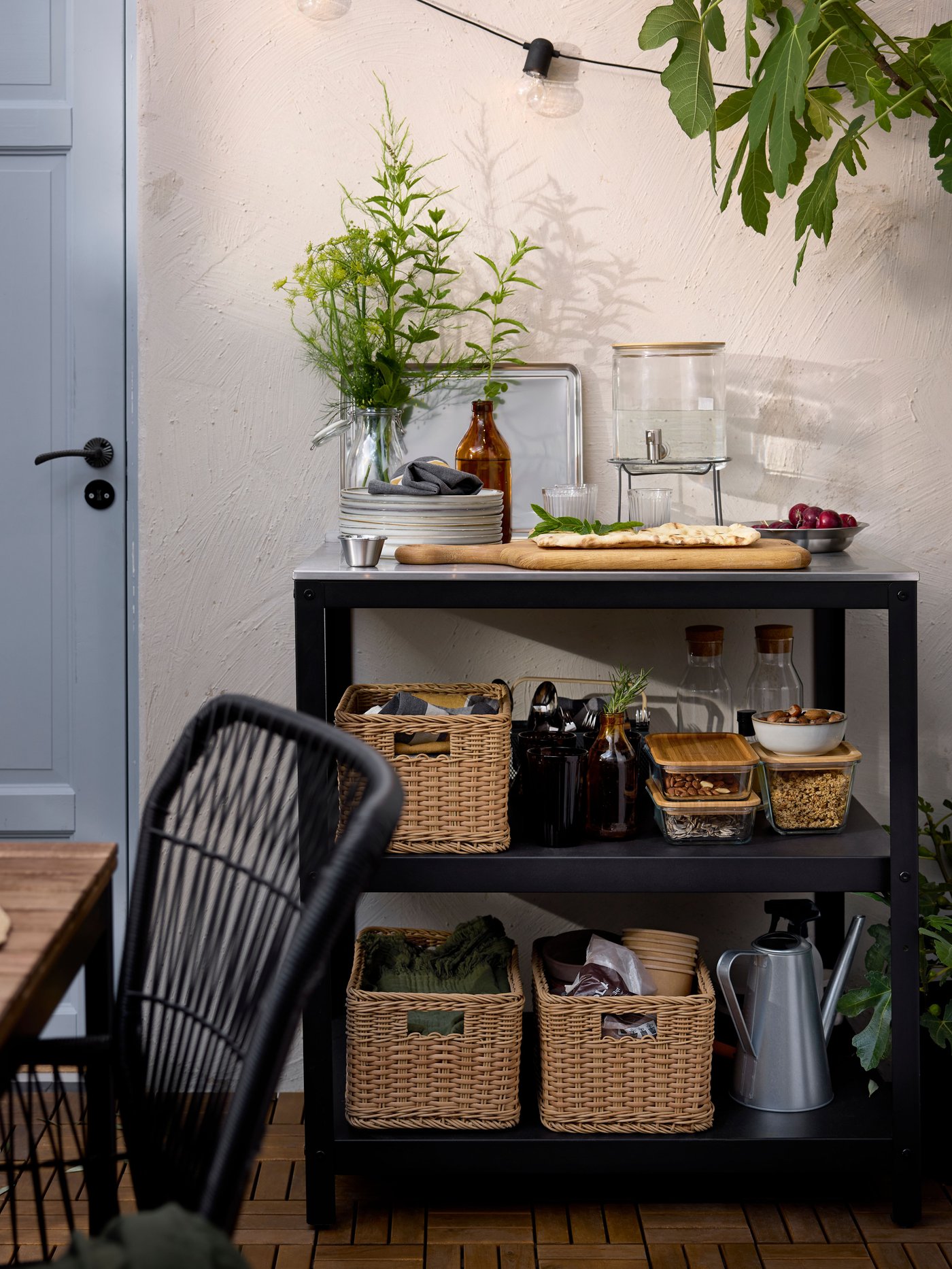 Un patio con una isla de cocina GRILLSKÄR de acero inoxidable con estantes abiertos llenos de comida y vajilla en cestas.