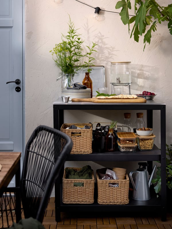 A patio with a GRILLSKÄR kitchen island in stainless steel with open shelves filled with food and dinnerware in baskets.