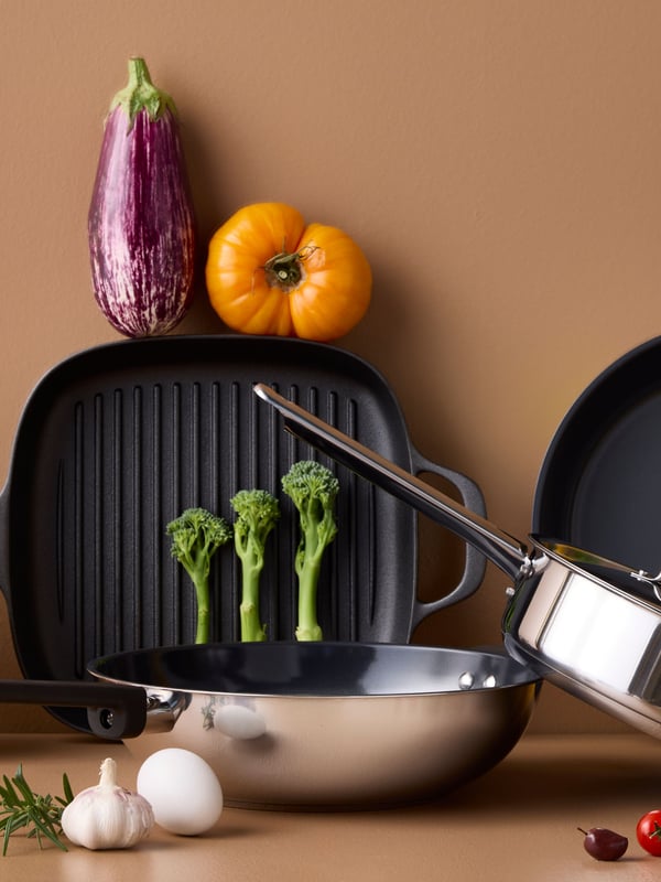 Four pans of different styles and materials are placed on a brown surface with different kinds of vegetables around them.