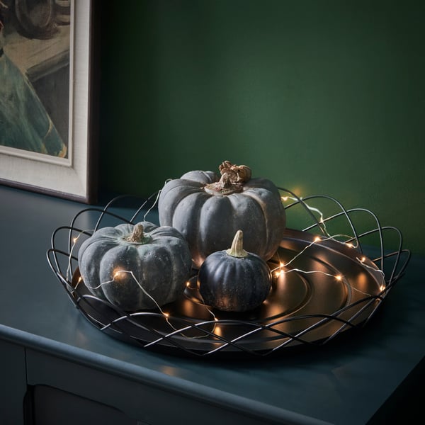 Su un buffet, un vassoio decorativo HÖSTAGILLE nero adornato da illuminazioni e zucche verdi.