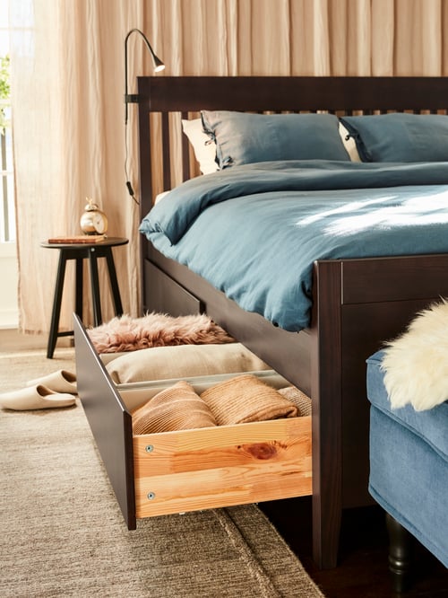 A bed frame with storage with one open drawer to the side, a round table, curtains, and an LED wall clamp spotlight.