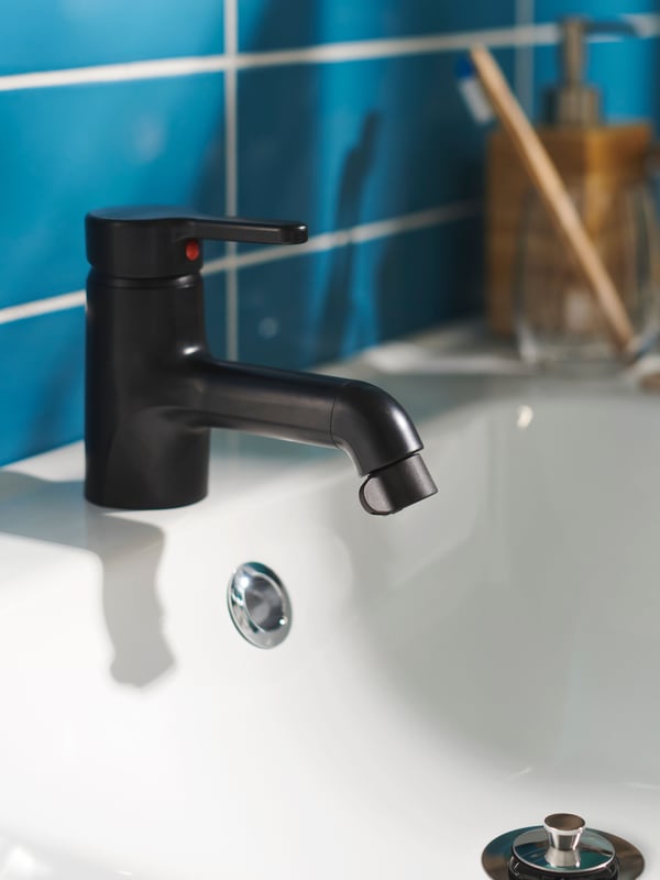 Eine schwarze, wassersparende ÅBÄCKEN Zerstäuberdüse, montiert auf einer schwarzen SÄLJAN Badarmatur, auf einem weissen Waschbecken mit blauen Wandfliesen dahinter.