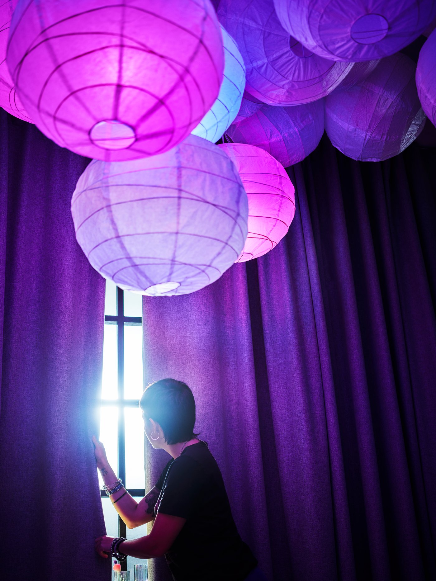Una donna sta aprendo delle tende oscuranti ROSENMANDEL. Al soffitto, ci sono dieci paralumi a sospensione REGOLIT illuminati con luci colorate.