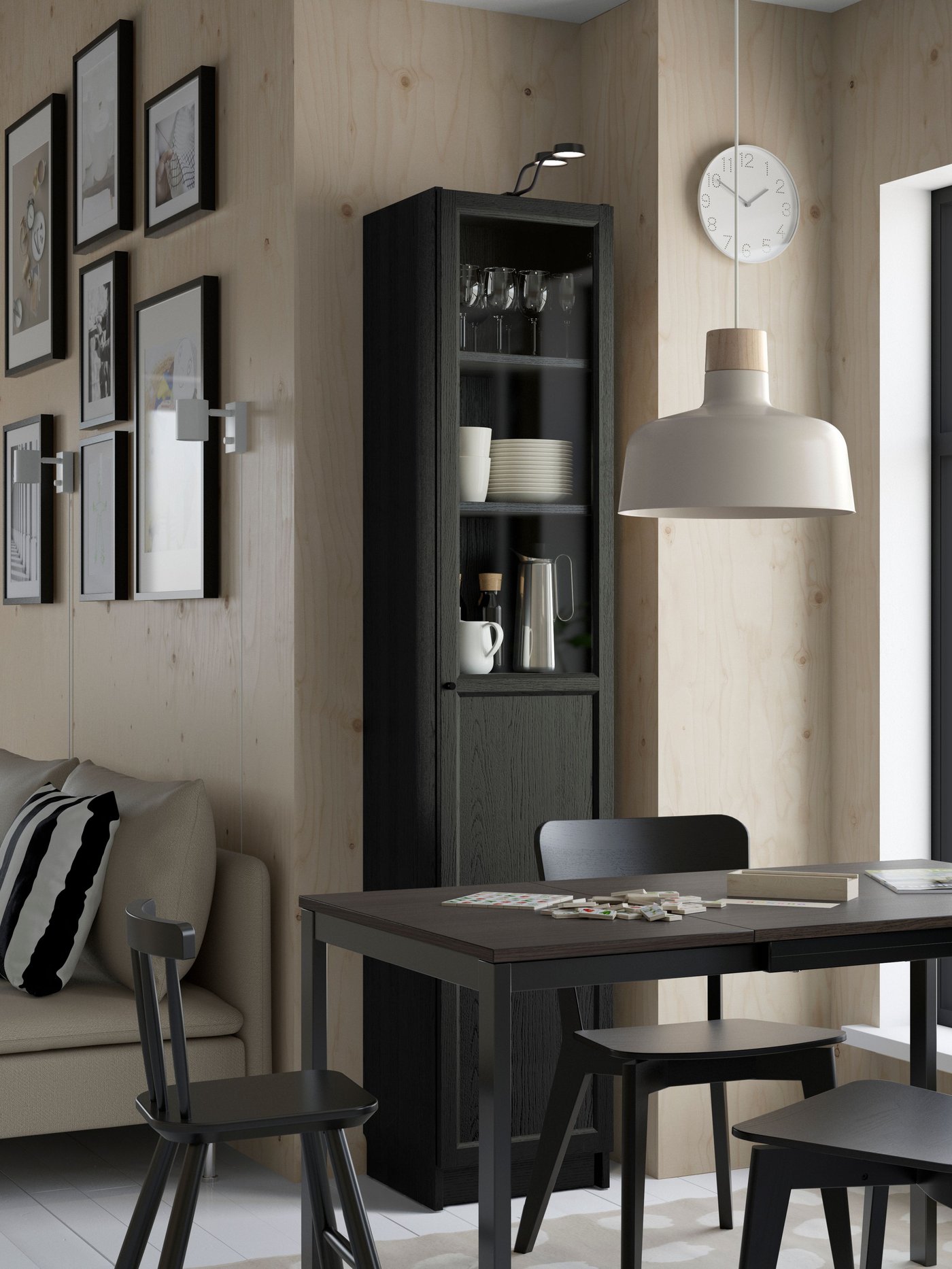 Matplats med svart matbord och stolar, högt vitrinskåp med glas och karaff, samt vit BUNKEFLO taklampa. Vägg med ljusa träpaneler och svartvita tavlor ger en modern känsla.
