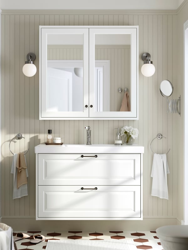 A white TÄNNFORSEN wash-stand with drawers and a mirror cabinet above it are placed in a bathroom with light brown walls.