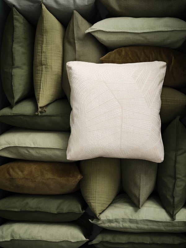 Un coussin habillé d’une housse JÄTTEGRAN blanc cassé repose sur une pile de coussins empilés en vert et brun.