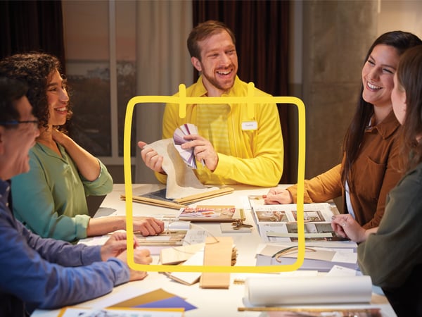 Smiling IKEA co-worker in a yellow sweater sitting at a table with four customers, reviewing materials, colour samples and planning documents.