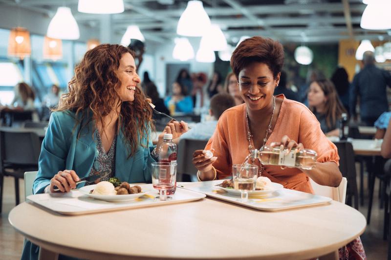 Deux jeunes femmes assises à la table ronde d'un restaurant et mangeant en souriant.