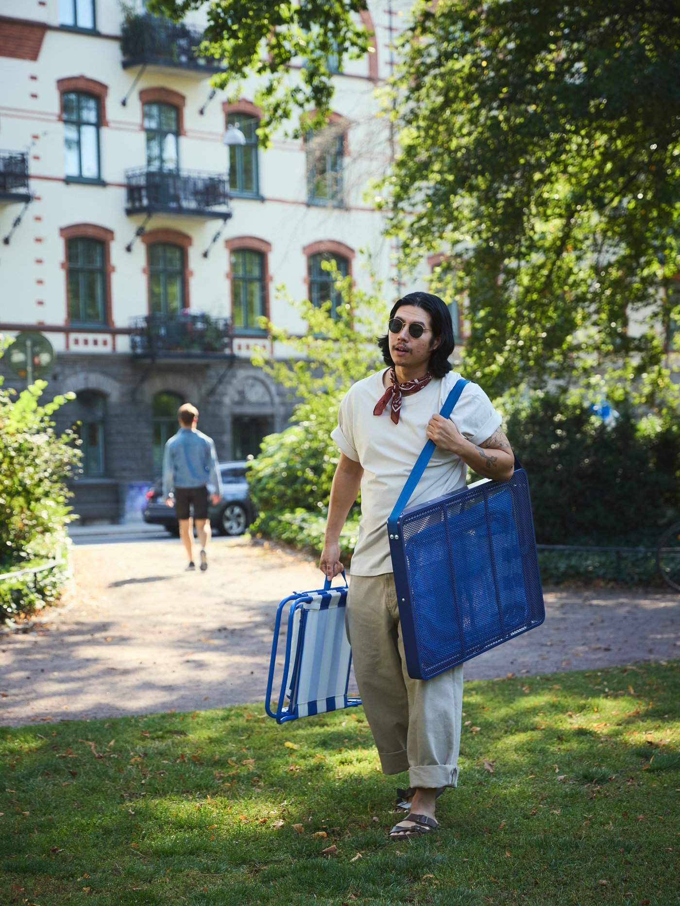 Ein Mann geht mit einem blauen STRANDÖN-Klapptisch und -Stuhl auf dem Weg zu einem Picknick durch den Park.