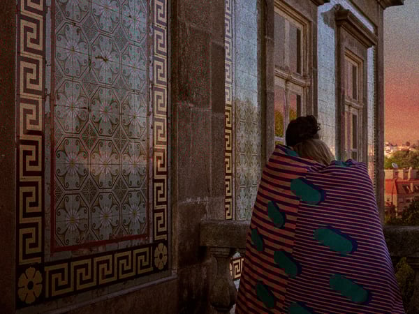 two individuals, seen from behind, embracing while looking out of a window or balcony. They are wrapped in a blanket with a distinctive green and blue pattern. The surrounding architecture is ornate, featuring intricate geometric patterns and floral motifs on the wall and floor tiles.