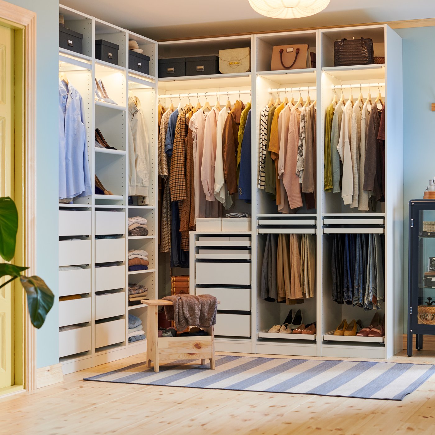 A pine PERJOHAN stool with storage stands in front of a large open PAX corner wardrobe with clothes, shoes, boxes and bags.