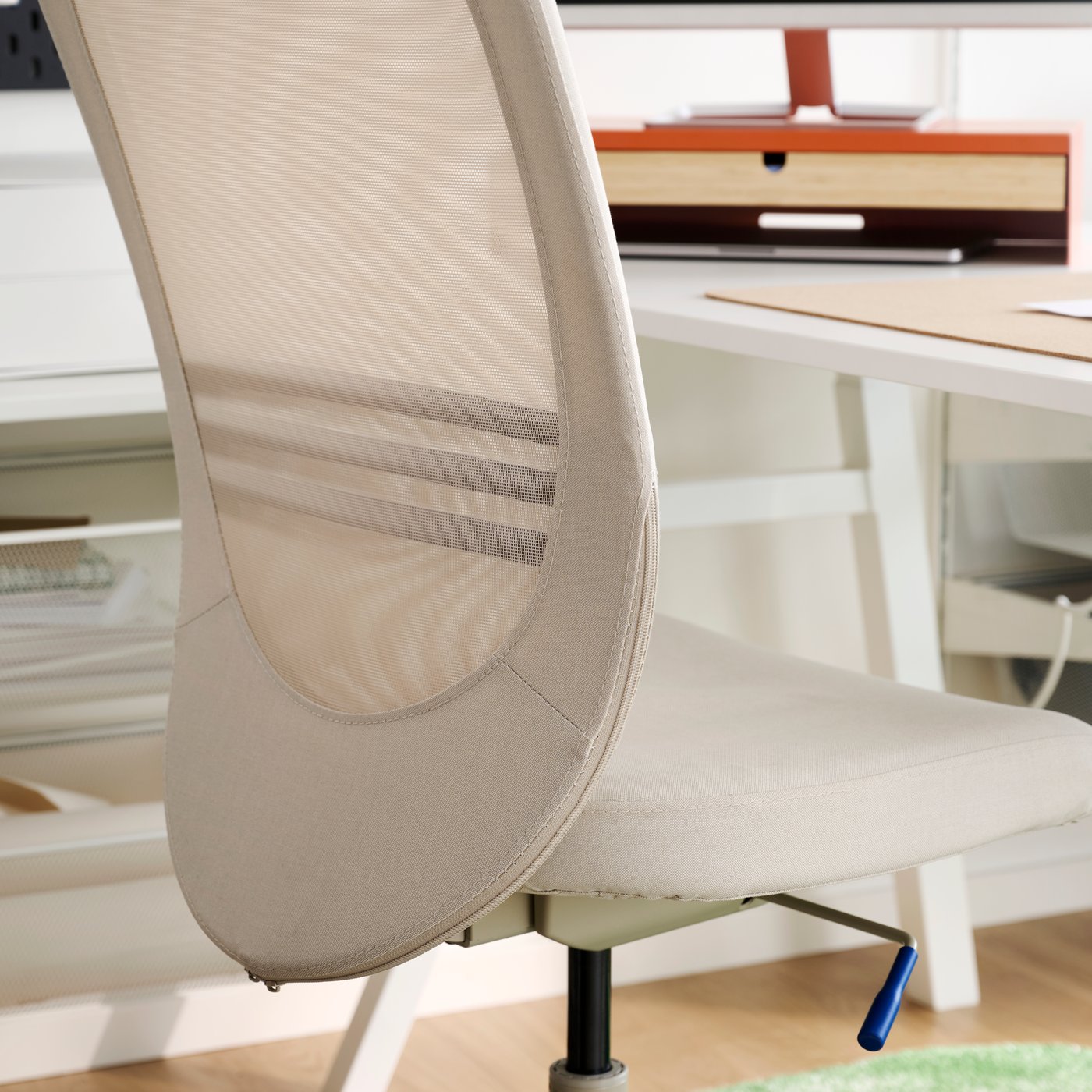 A close-up of a beige FLINTAN office chair with a mesh backrest sits in a bright office space near a desk.