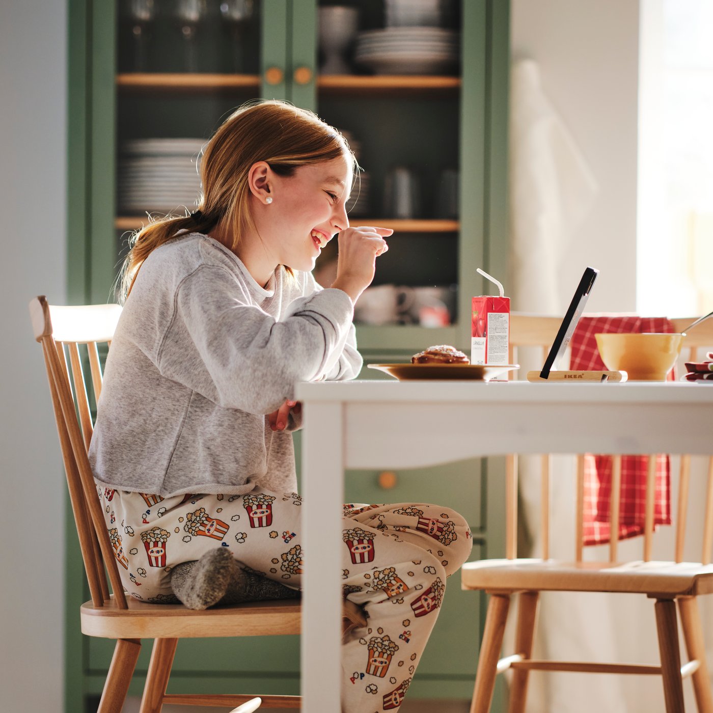 A little girl sits at the dining table enjoying a cinnamon bun and a juice while face timing with someone on her tablet.