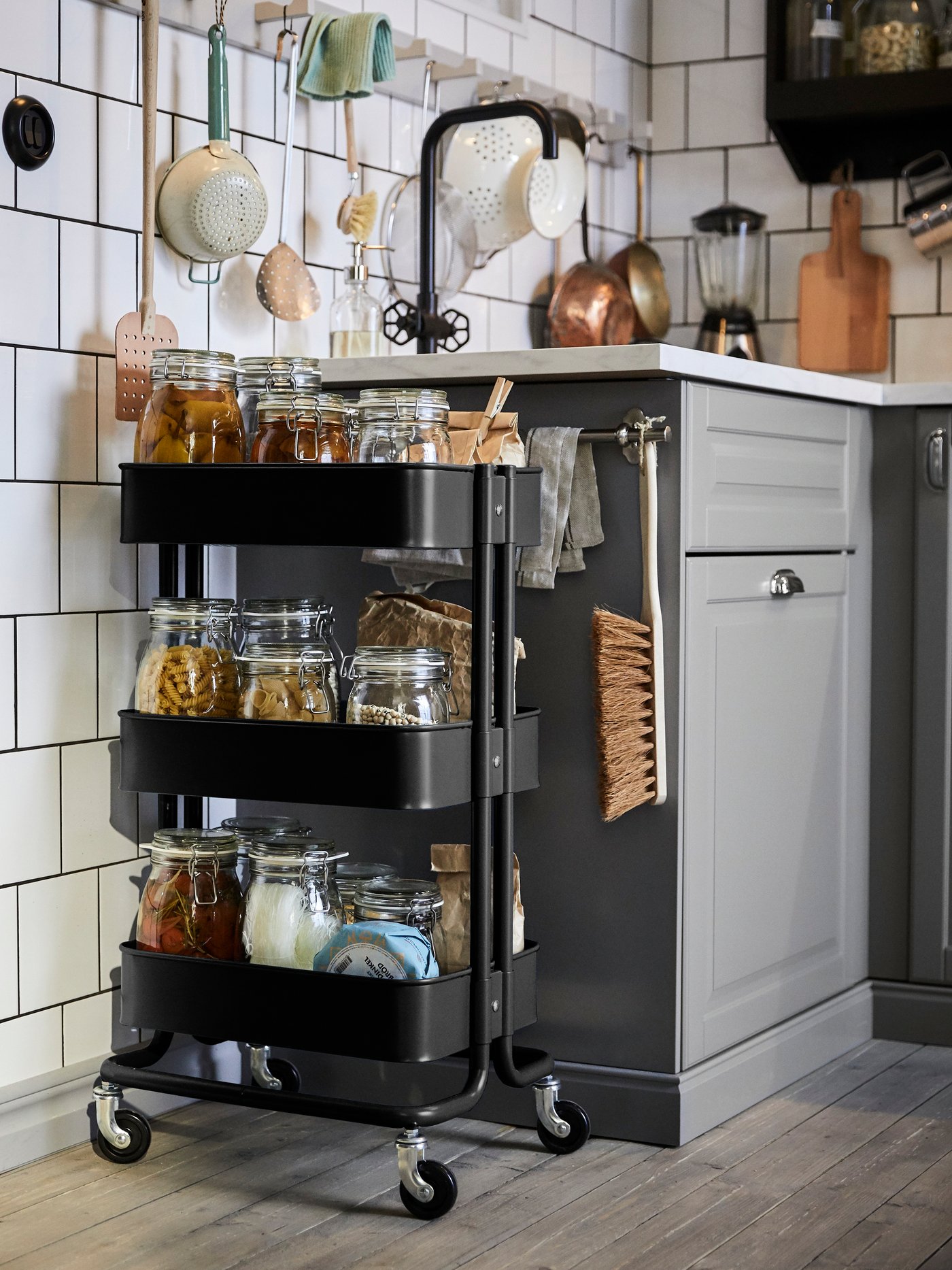 Ein schwarzer RÅSKOG Wagen mit Glasbehältern voller verschiedener Lebensmittel auf den Regalböden steht neben einem Spülbecken in einer weiß gefliesten Küche.