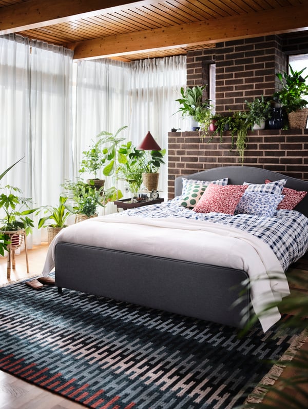 A bedroom with plants, a red lamp, a grey HAUGA upholstered bed frame, a blue and white quilt cover, and six cushions.