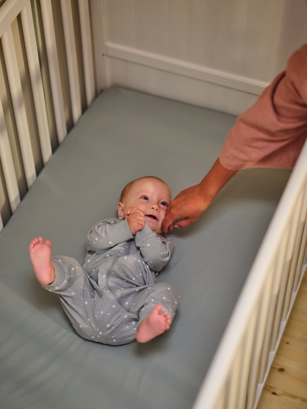 A baby lies in a SUNDVIK cot that has a blue GULDVÄVARE sheet on the mattress, with an adult’s hand stroking its cheek.