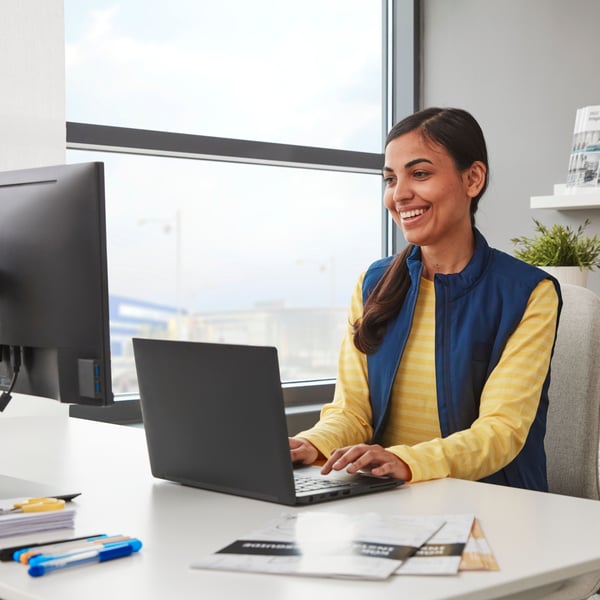 A smiling person in a yellow sweater and a blue gilet is working on a black laptop, which is resting on a white desk next to a window.