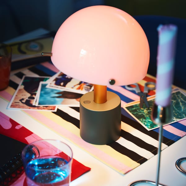 A light pink/dark grey-green, battery operated NÖDMAST LED portable lamp placed on colourful placemats next to a blue glass.