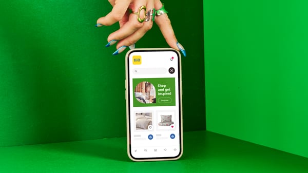 A hand with fingernails painted green is holding a smartphone displaying the IKEA app against a green background.
