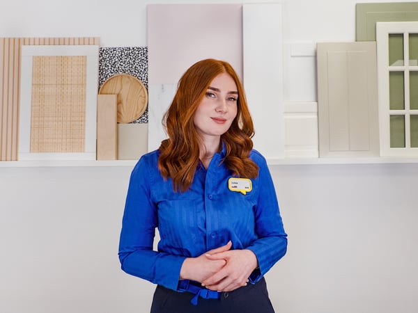 Projektantka IKEA stojąca w niebieskiej koszuli na tle ściany z półką, na której ustawione są próbki materiałów i fronty kuchenne.